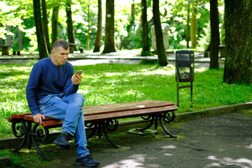 Young man sitting on a bench in a park and holding mobile phone. Lifestyle Outdoor street photo