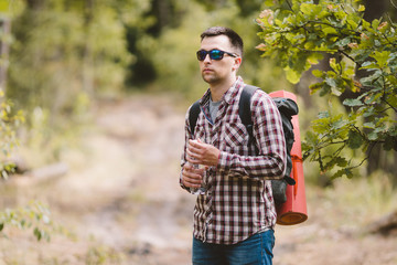 Fototapeta premium Hiker Drinking Water In Forest. Tired man drinking water from bottle in woodland. Handsome Traveler With Backpack And Flask In Forest. vacation, outdoor lifestyle freedom concept. Adventures hiking