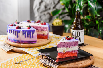 Beautiful layered berry cake with a purple, white and pink layer, decorated with raspberries and blueberries on top. Beautiful cake in a cut on a black saucer on a wooden table in the restaurant