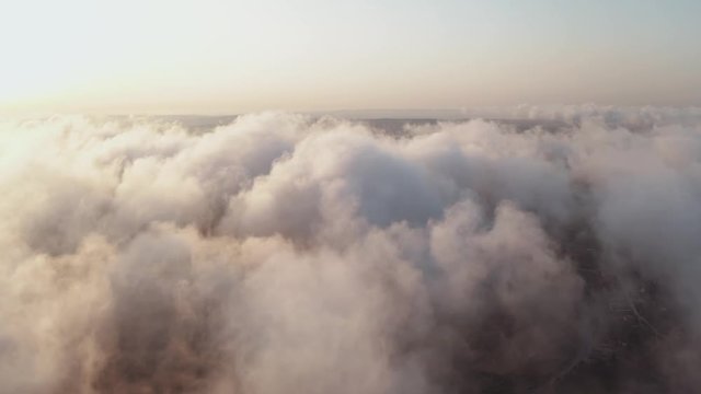 Scenic aerial view of moving white clouds. The drone flies high back in the blue sky through the fluffy clouds in the evening at the bright sun. The sun is hidden behind the clouds at sunset fog