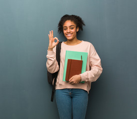 Young student black woman cheerful and confident doing ok gesture. She is holding books.