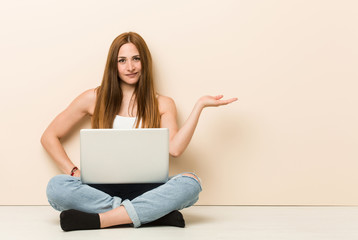 Young ginger woman sitting on her house floor showing a copy space on a palm and holding another hand on waist.