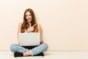 Young ginger woman sitting on her house floor smiling and pointing aside, showing something at blank space.