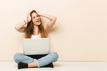 Young ginger woman sitting on her house floor laughs joyfully keeping hands on head. Happiness concept.