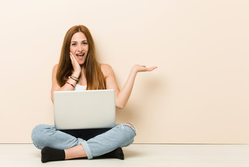 Young ginger woman sitting on her house floor holds copy space on a palm, keep hand over cheek. Amazed and delighted.