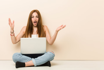 Young ginger woman sitting on her house floor receiving a pleasant surprise, excited and raising hands.