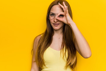 Young redhead ginger woman with freckless excited keeping ok gesture on eye.