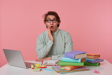 Studio shot of young wondered guy with glasses, wears on blank shirt, sitting at a table with books, working at a laptop, looks surprised and shocked. Isolated over pink background.