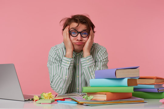 Young Tired Man With Glasses,sitting At A Table With Books, Working At A Laptop, Looks Unhappy And Sad, Sadly Looks Up And Dreams Of Leaving Home As Soon As Possible, Isolated Over Pink Background.