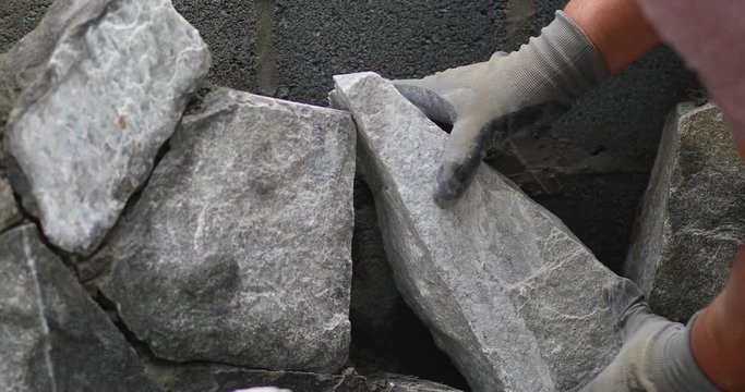 A man working in construction wears protective gloves and places a heavy rock on top of a wall of rocks in a process to build an outdoor fireplace.