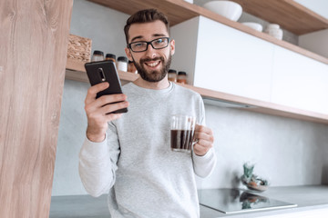 smiling man with smartphone and Cup of coffee standing in his kitchen