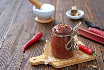 Spicy rhubarb chutney sauce in a glass jar on a wooden background. Preserving, harvesting. Indian food. - obrazy, fototapety, plakaty
