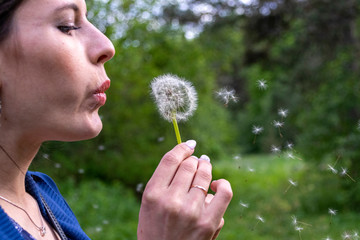 Happy beautiful woman blowing dandelion over sky background, having fun and playing outdoor, teen girl enjoying nature, summer vacation and holidays, young pretty female holding flower, wish concept