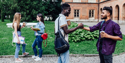 Fototapeta premium Meet of students. Two handsome young male student holding coffe, have break time while walking on green grass near university.