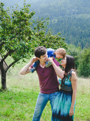 Happy young family and little son playing and having fun outdoors. Green mountains on background. Lifestyle. Sincere moments of family. Child sits on the shoulders of his father. Leisure, childhood.