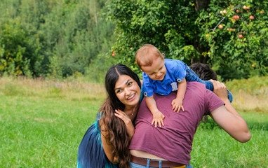 Naklejka premium Happy young family and little son playing and having fun outdoors. Green mountains on background. Lifestyle. Sincere moments of family. Child sits on the shoulders of his father. Leisure, childhood, p