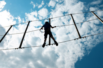 Boy climbing rope trail in an adventure park. Silhouettes in front of a cloudy sky background.a