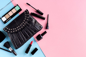 Black leather bag with nail polish, pomade, mascara and eye shadows on pink and blue background composition.