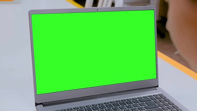 Woman sitting at white table and looking at grey laptop computer device with blank green screen in cafe, home or office. Technology, chroma key, template, mock up and entertainment concept