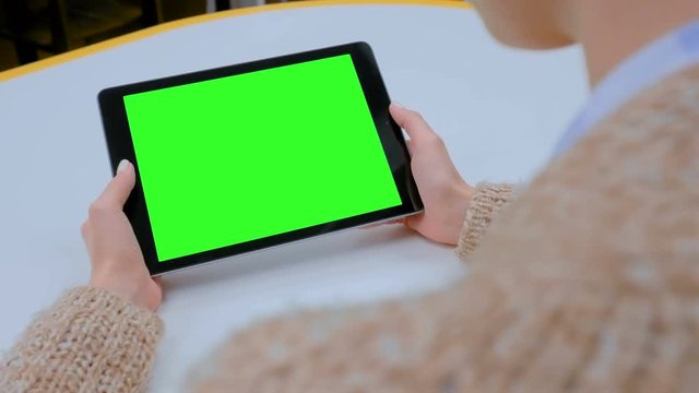 Over shoulder view - woman looking at black digital tablet computer device with empty green screen on table in cafe, home or office. Technology, chroma key, template, mock up, entertainment concept