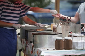 A French chef, in blue and white striped shirt, is preparing crepes or galettes on a hot-plate at a market stall. Another stall holder is giving change to a customer in the background. 