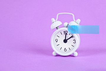 White alarm clock violet pastel background. Blank sticky note on clock. Space copy. Minimal concept.