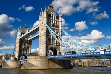 Fototapeta na wymiar Twin stone towers of Tower Bridge over the Thames River in London with red bus and blue and white suspension bridge and elevated pedestrain walkway