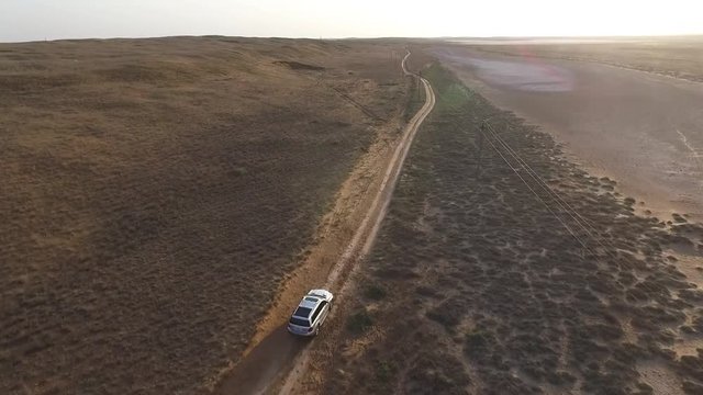 Back aerial view: Big silver SUV driving in a steppe in front of the sunset