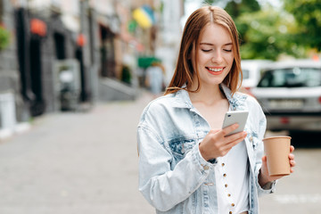 Fototapeta premium Happy young woman standing and holding a smartphone looking at the screen outdoor- Image