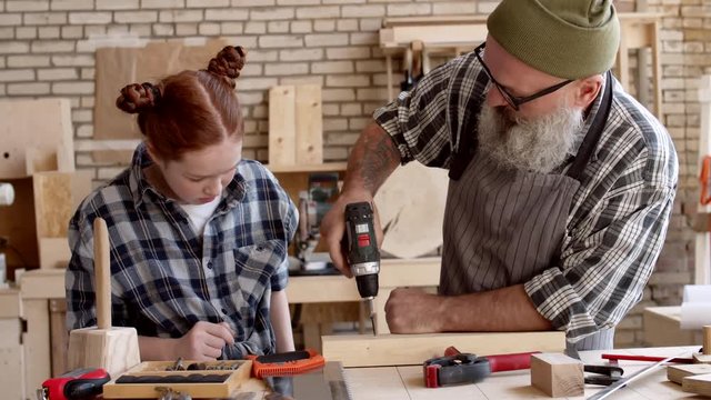 Lockdown of Caucasian aged hipster man showing his granddaughter with red hair how to use electrical screwdriver in carpentry shop