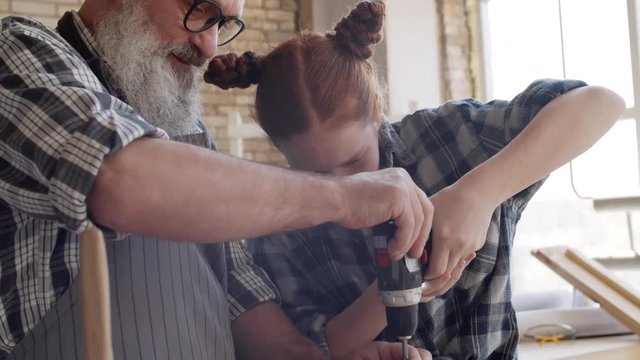 Medium shot of Caucasian aged hipster man helping his granddaughter with red hair screwing screw in timber using electrical screwdriver in carpentry shop