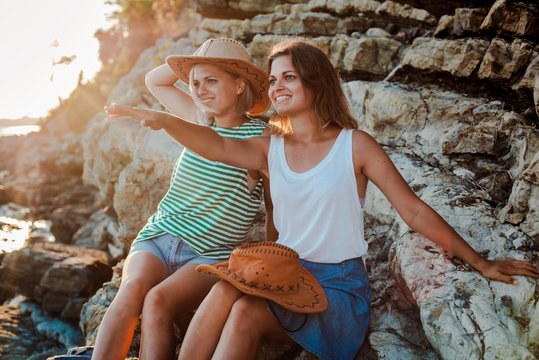 Two young cheerful women in hipsters hats on a rock on the coast of the sea. Summer landscape with girl, sea, Islands and orange sunlight.