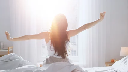 Foto op Aluminium Beautiful Brunette is Waking up in the Morning, Stretches in the Bed, Sun Shines on Her From the Big Window. Happy Young Girl Greets New Day with Warm Sunlight Flare. © Gorodenkoff