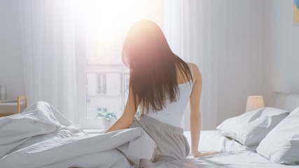 Fototapeta na wymiar Beautiful Brunette is Waking up in the Morning, Stretches in the Bed, Sun Shines on Her From the Big Window. Happy Young Girl Greets New Day with Warm Sunlight Flare.
