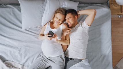 Obraz premium Loving Young Couple Spending Morning in Bed, Pregnant Young Woman Shows Her Partner Something on a Touchscreen Smartphone. Searching for Baby Clothes, Browsing Through Social Media, Posting Pictures.