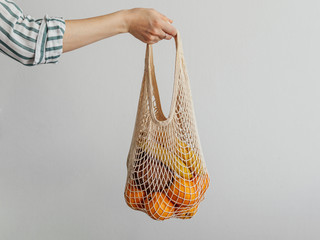 Mesh bag with fruits in female hand. Stylish young woman hand hold mesh shopping bag on light gray...