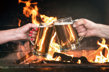 People are holding in hands a glasses with a beer and are clinking on a burning fire background.