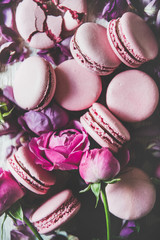 Sweet pink macaron cookies and spring rose flowers, buds and petals over wooden background, top view, selective focus, close-up. Food texture, background and wallpaper