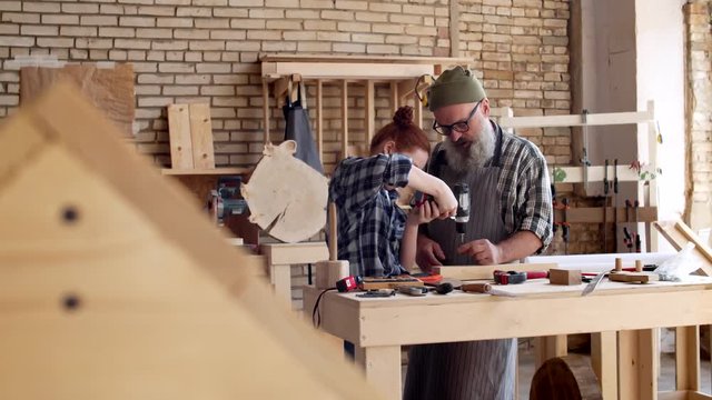 Medium shot of Caucasian aged hipster man and his granddaughter with red hair using electrical screwdriver in carpentry shop