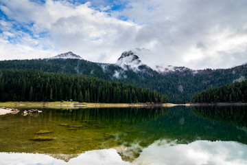 Fototapeta na wymiar Montenegro, Reflecting calm waters of black lake in durmitor national park surrounded by green forest and high snow covered mountains partly hidden in fog near zabljak