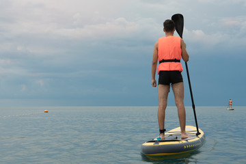 Young man on sup board in a life jacket on the background of the sea. View from back surfer