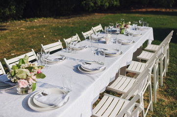 Fototapeta na wymiar A large white long table with chairs, decorated with fresh flowers, with plates, glasses and forks, stands in a park with green grass. Wedding decorations and details. Preparing for a wedding party.