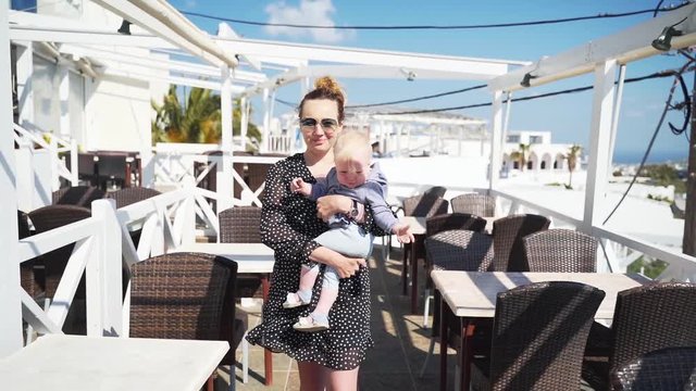 Young mother with child in her arms goes on the terrace in a restaurant overlooking the sea, Santorini, Greece. Hiking with baby. Slow motion