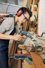 Professional female carpenter in ear muffs and protective goggles cuttig wooden plank with circular...