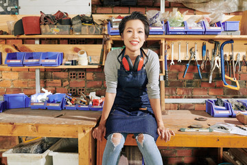 Happy pretty young Vietnamese female carpenter sitting on workbench in furniture workshop