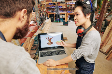 Vietnamese female carpenter showing 3D model of wooden table stand on laptop screen to coworker