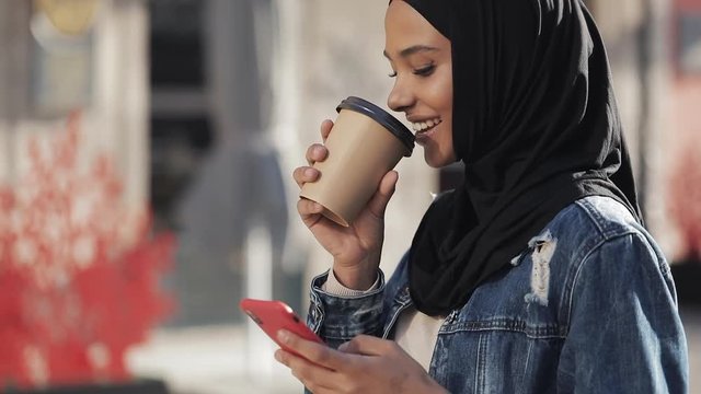 Smilng muslim woman wearing hijab headscarf walking in the city center, using smartphone and drinking coffee. Communication, online shopping, social network concept.