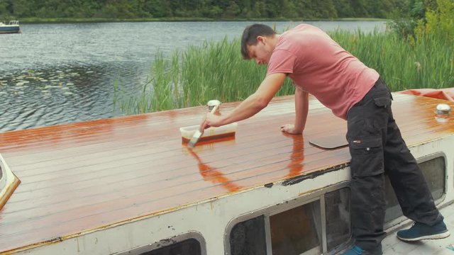Carpenter varnishes roof of wooden boat with brush on lake
