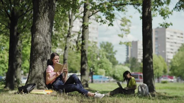 Beautiful female student sitting under tree leaning on trunk and reading book in park. Another college girl typing on laptop in background