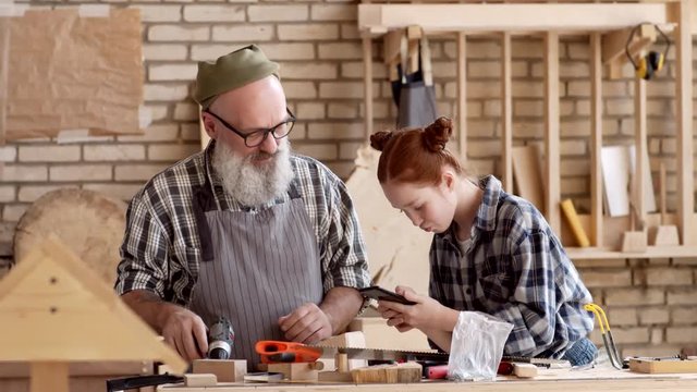 Medium shot of Caucasian ginger teenage girl standing in carpentry shop with telephone in her hands and shooting video with her hipster granddad working with wood using electrical screwdriver